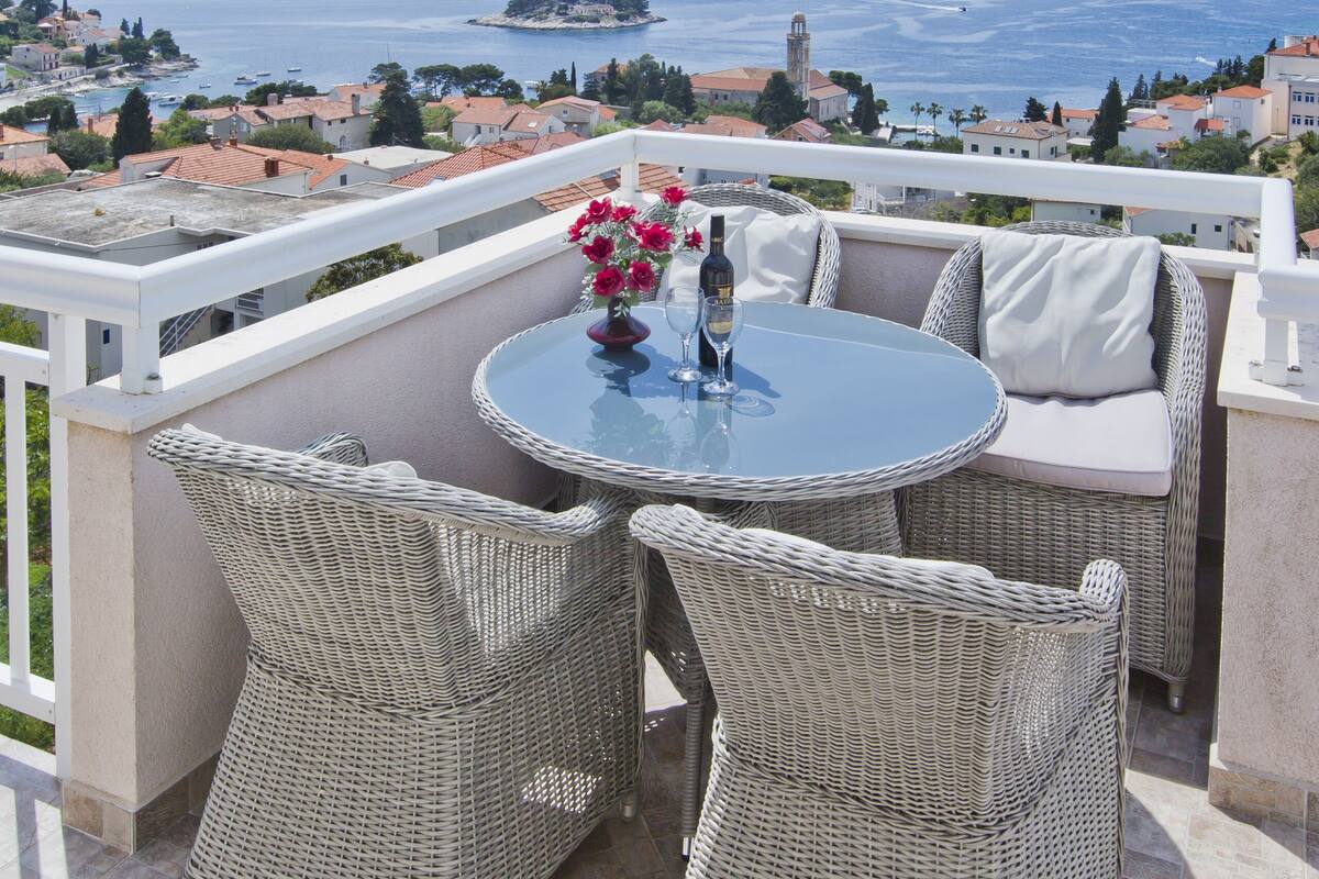 Un balcón con mobiliario de mimbre, una mesa con un jarrón de flores y vistas al mar y edificios a lo lejos.