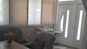 A modern living room with a grey armchair, a small table with a vase of flowers, and a large window with blinds. - Apartmani Kresic 1 (Hvar)