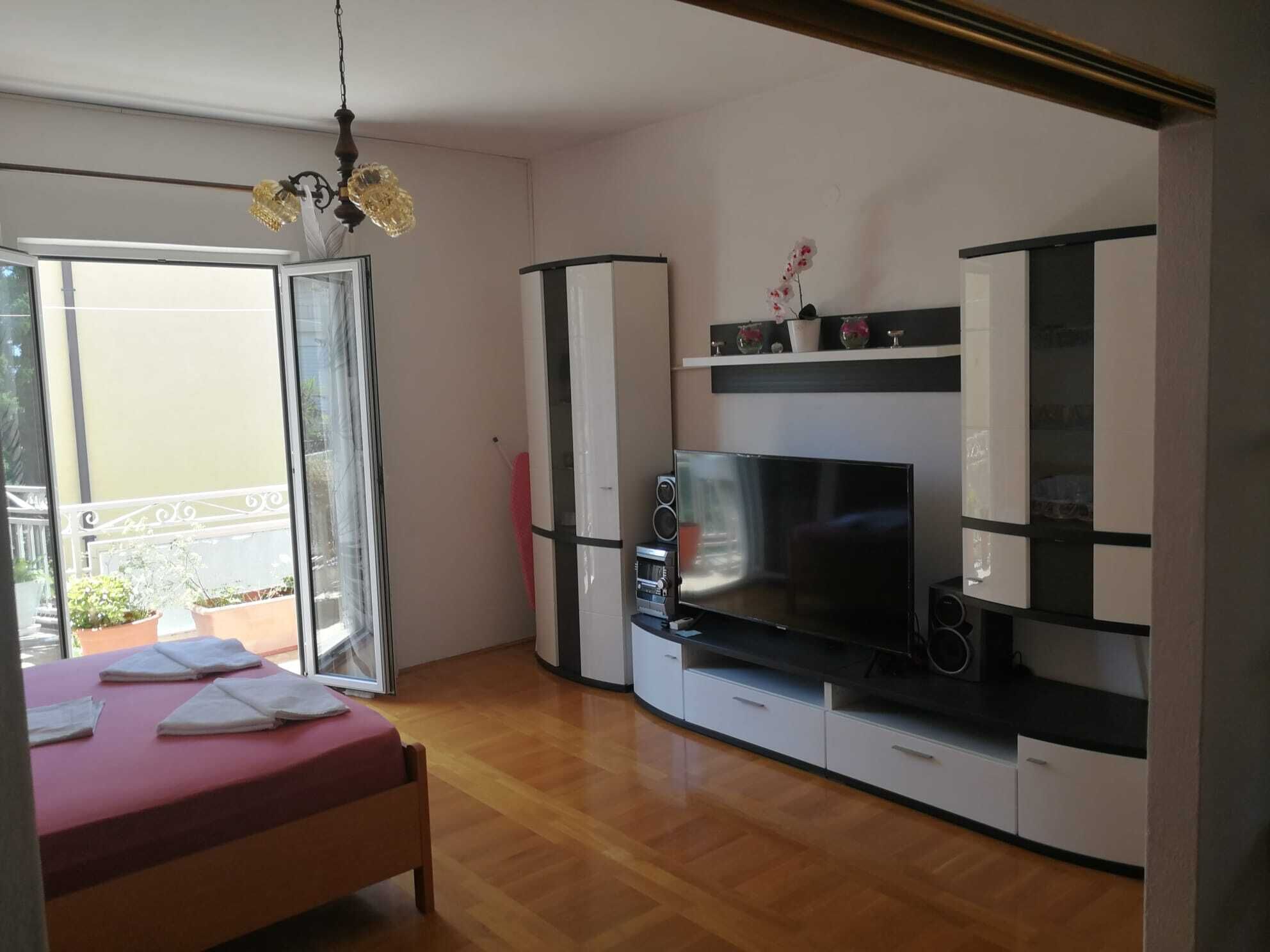 Un dormitorio con cama, televisor, una mesita, estante con adornos y puerta corrediza de vidrio que da a un balcón.