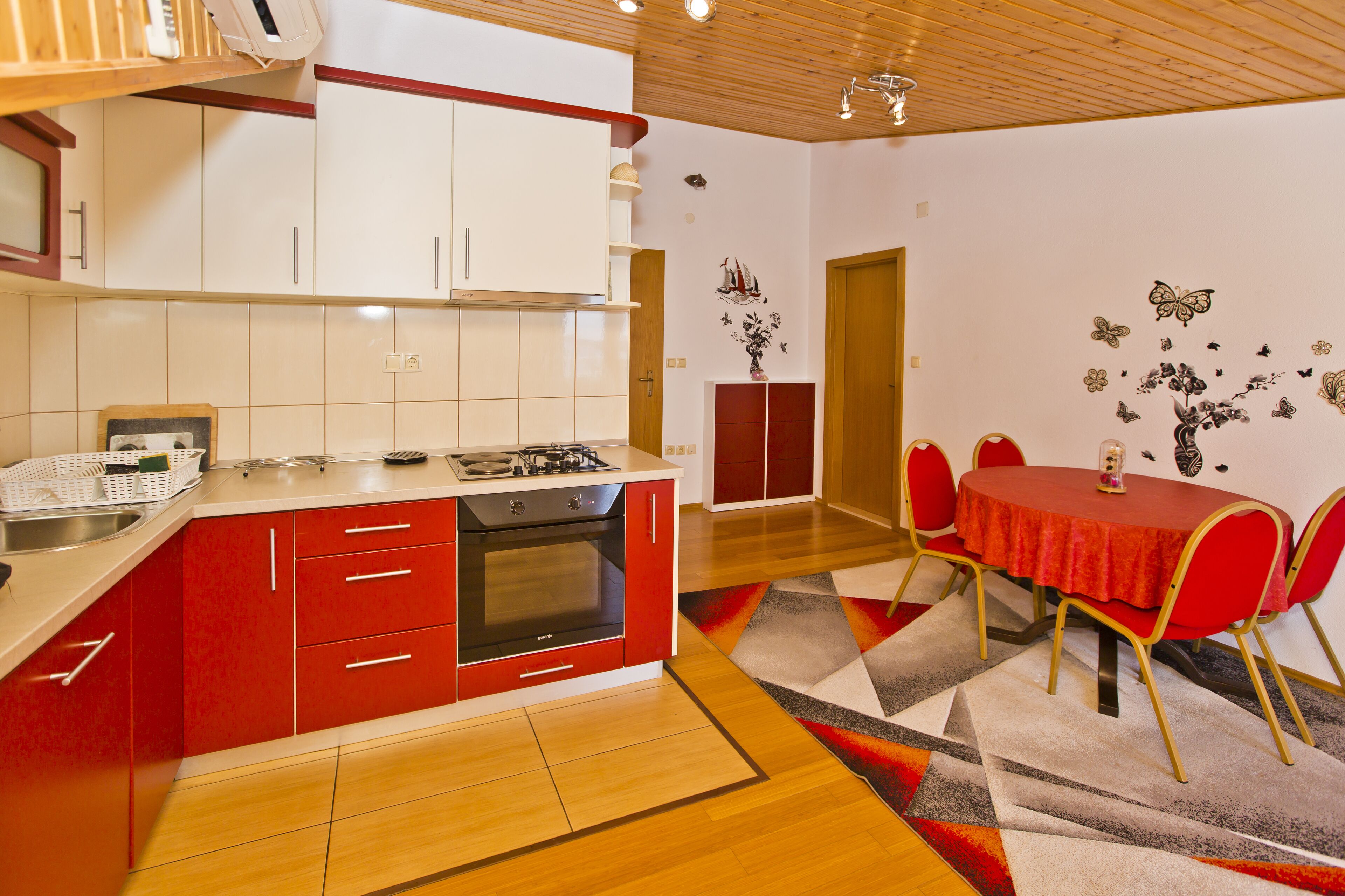 A compact kitchen with red and white cabinets, a built-in oven, and a dining area with a red table and chairs.