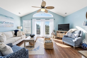 A living room with a high ceiling, a ceiling fan, a large window, a television, and a sofa. - Island Time: Golf Community, Beach Club Access w/ Pool and Oceanfront Parking (Nags Head)