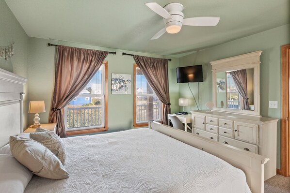 A bedroom with a bed, a desk, a dresser, a ceiling fan, and a TV on the wall.