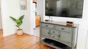 A living room with a flat-screen TV mounted on a wooden cabinet, a potted plant, and a kitchen area in the background. - Cozy Home in a Quiet Neighborhood

 (Vallejo)
