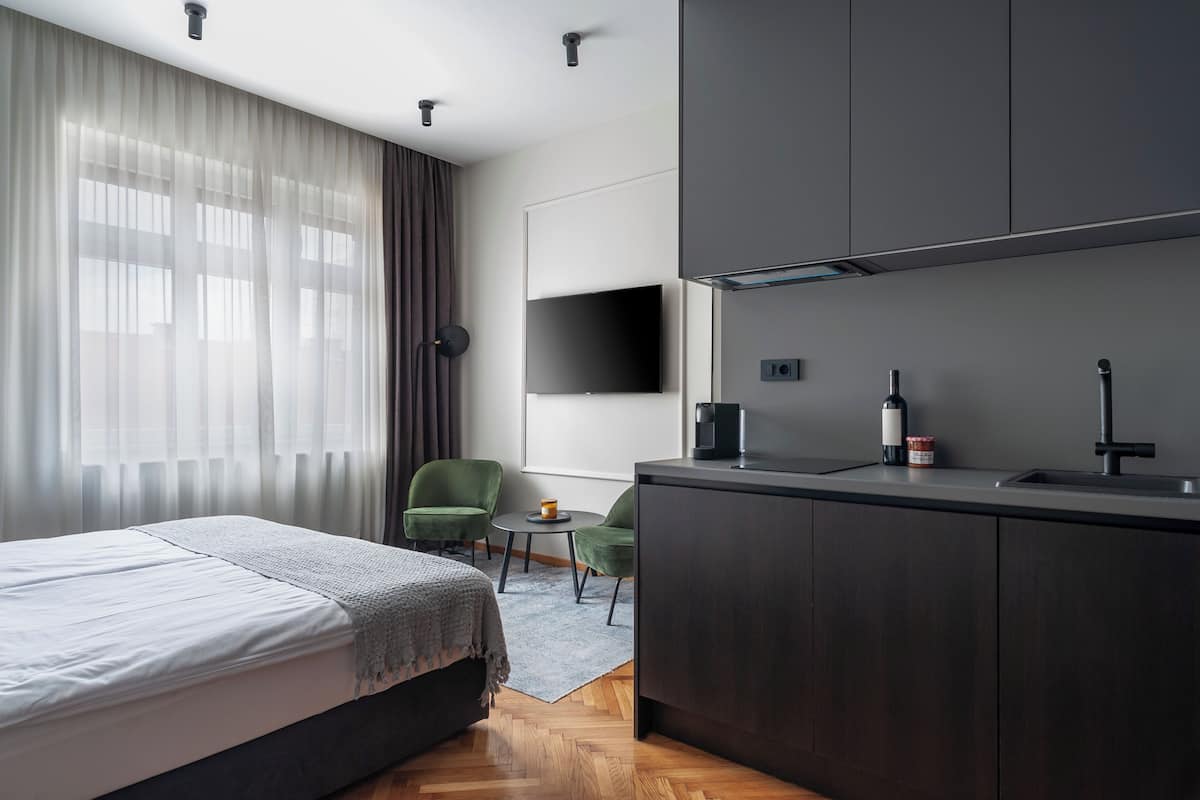 A modern hotel room with a bed, a small round table, two green chairs, a dark wooden desk, a flat-screen TV mounted on the wall, and a kitchen area with a sink and a wine bottle.