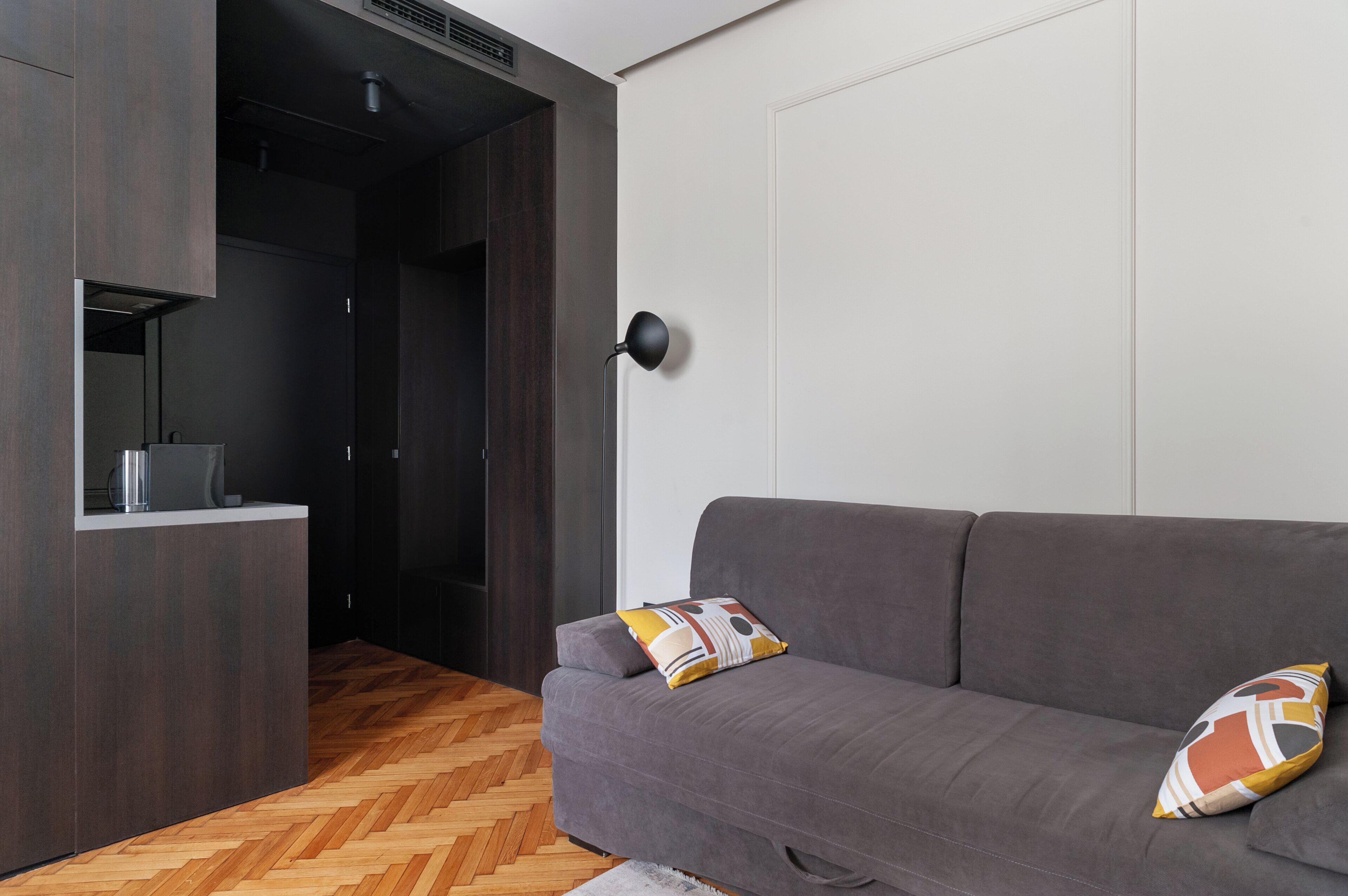 Un salón moderno con un sofá gris, dos cojines estampados, una lámpara de pie negra y un suelo de madera en espiga.