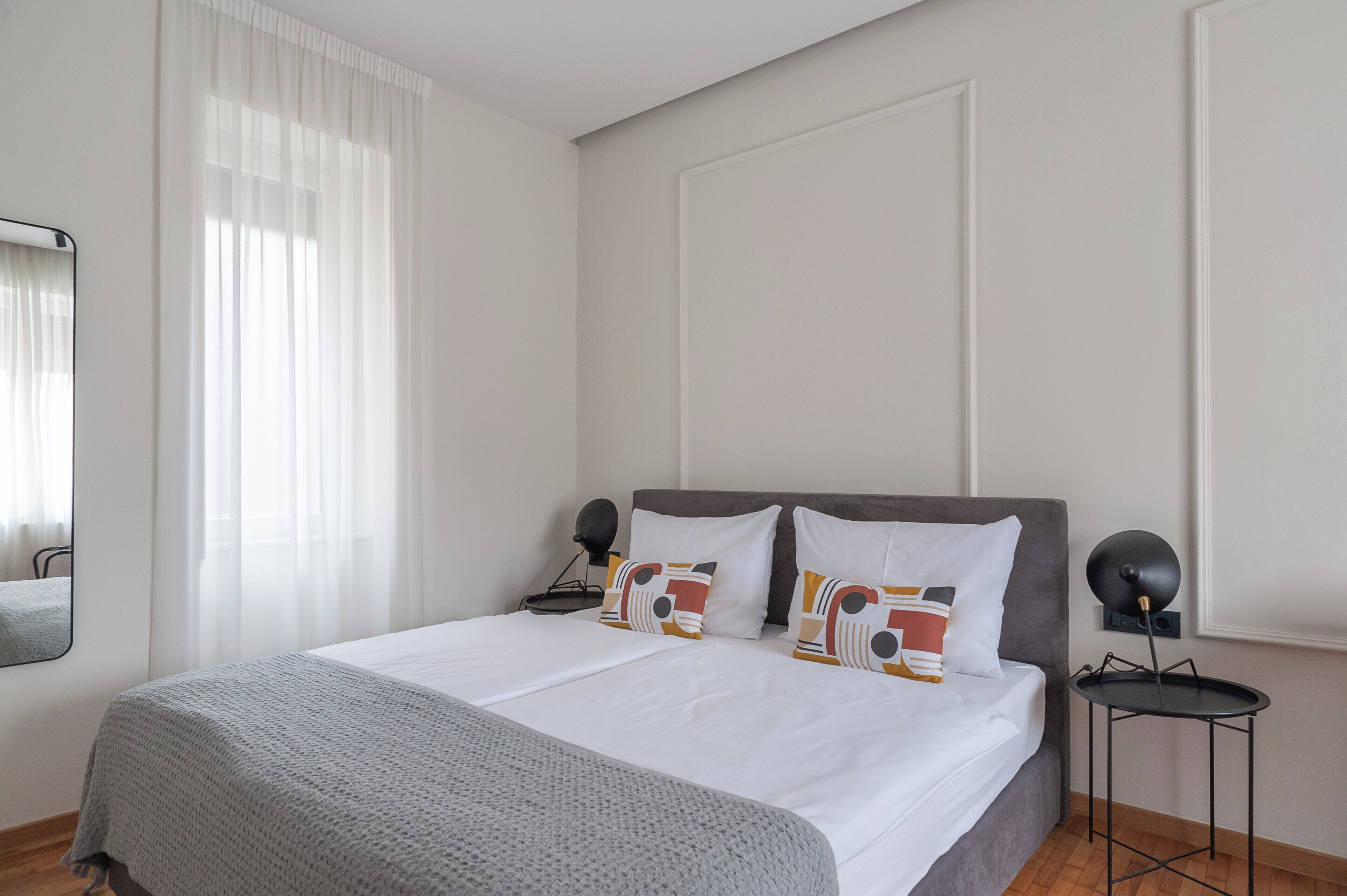 A neatly made bed with white linens and patterned pillows, a bedside table with a lamp, and a window with sheer curtains.