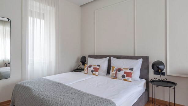 Una cama bien tendida con sábanas blancas y almohadas estampadas, una mesita de noche con lámpara, y una ventana con cortinas de encaje.
