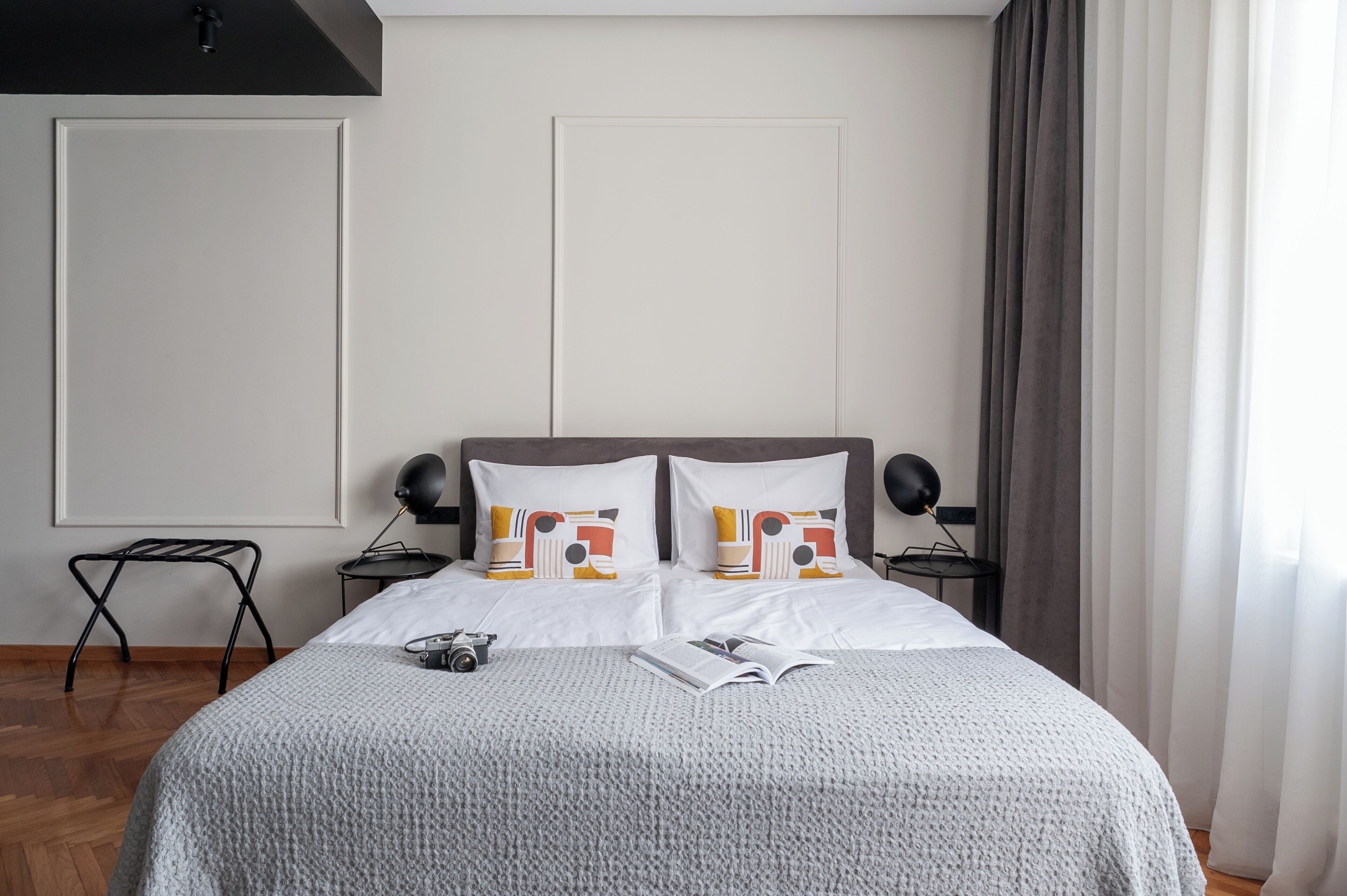 Una cama bien hecha con un edredón gris, dos almohadas con un estampado colorido, una mesita de noche con una lámpara y una pequeña mesa con una cámara.
