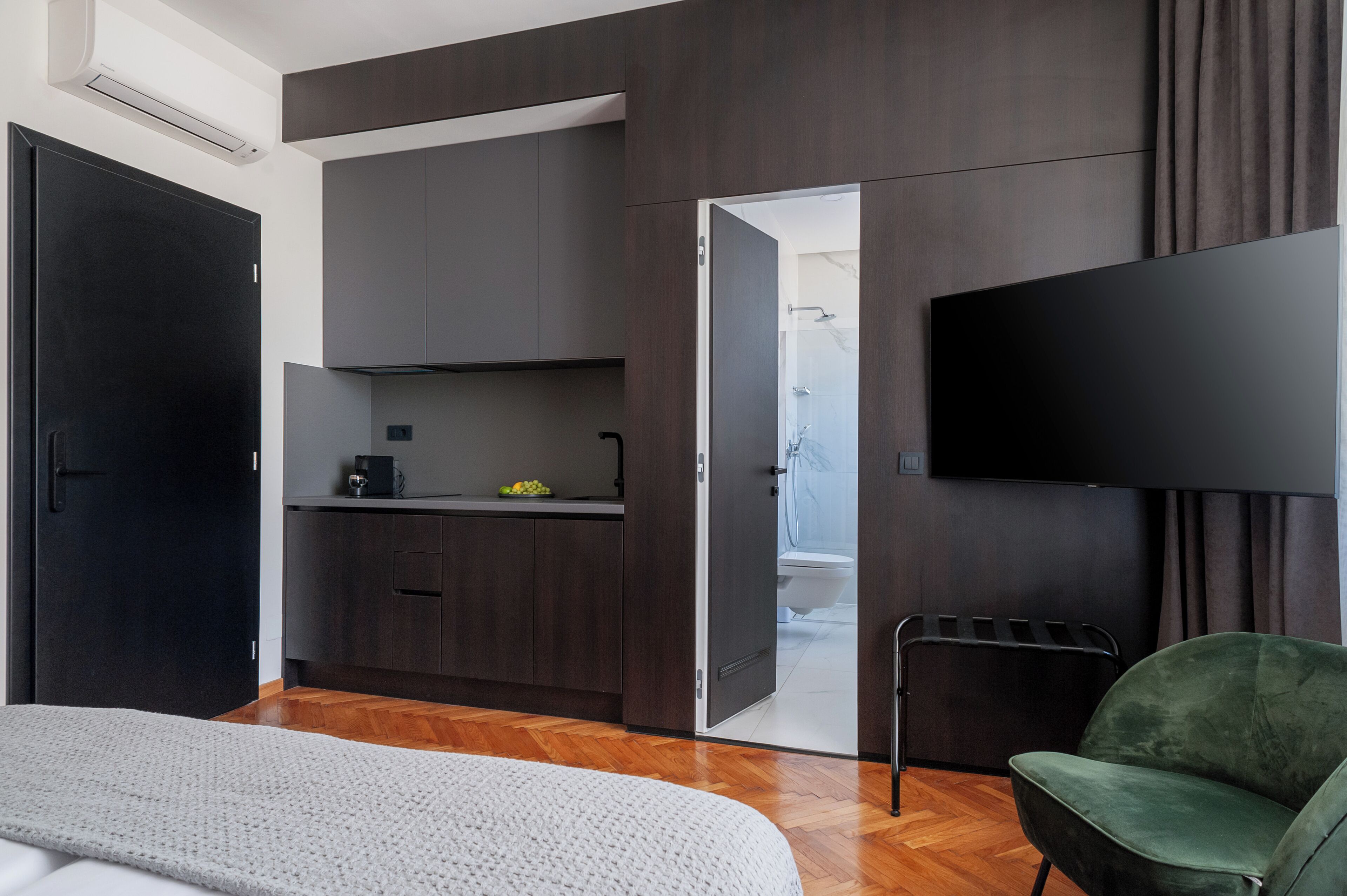 A modern room with a bed, a green chair, a flat-screen TV, a kitchenette, and a bathroom visible through an open door.