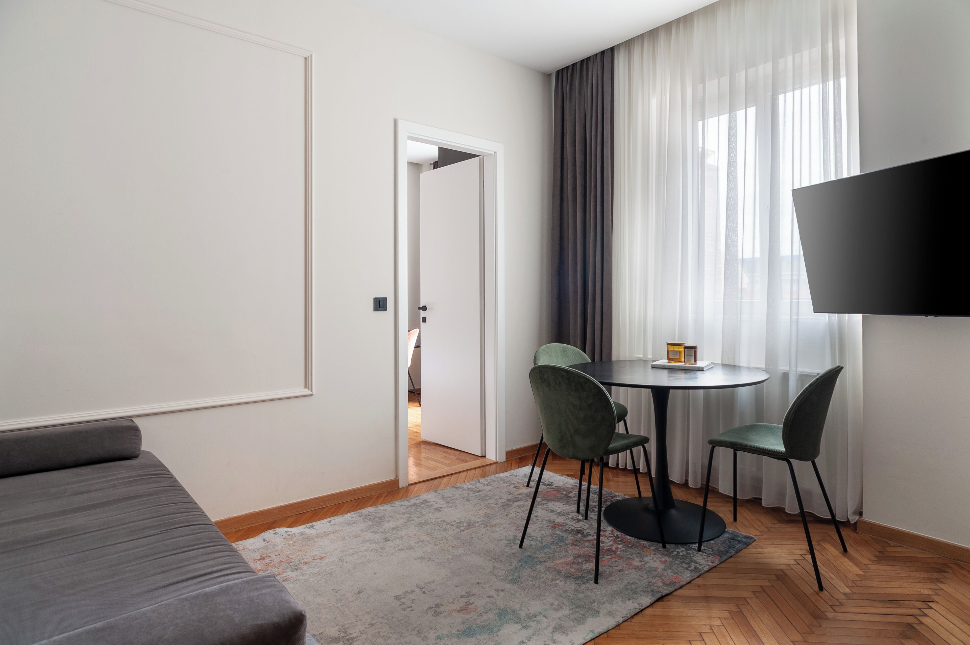 Una habitación de hotel moderna con sofá, una mesa de comedor con sillas, televisión y vistas al exterior a través de cortinas translúcidas.