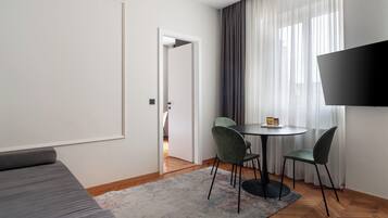 Una habitación de hotel moderna con sofá, una mesa de comedor con sillas, televisión y vistas al exterior a través de cortinas translúcidas.