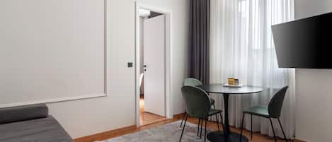 A modern hotel room with a sofa, a dining table with chairs, a TV, and a view of the outdoors through sheer curtains.