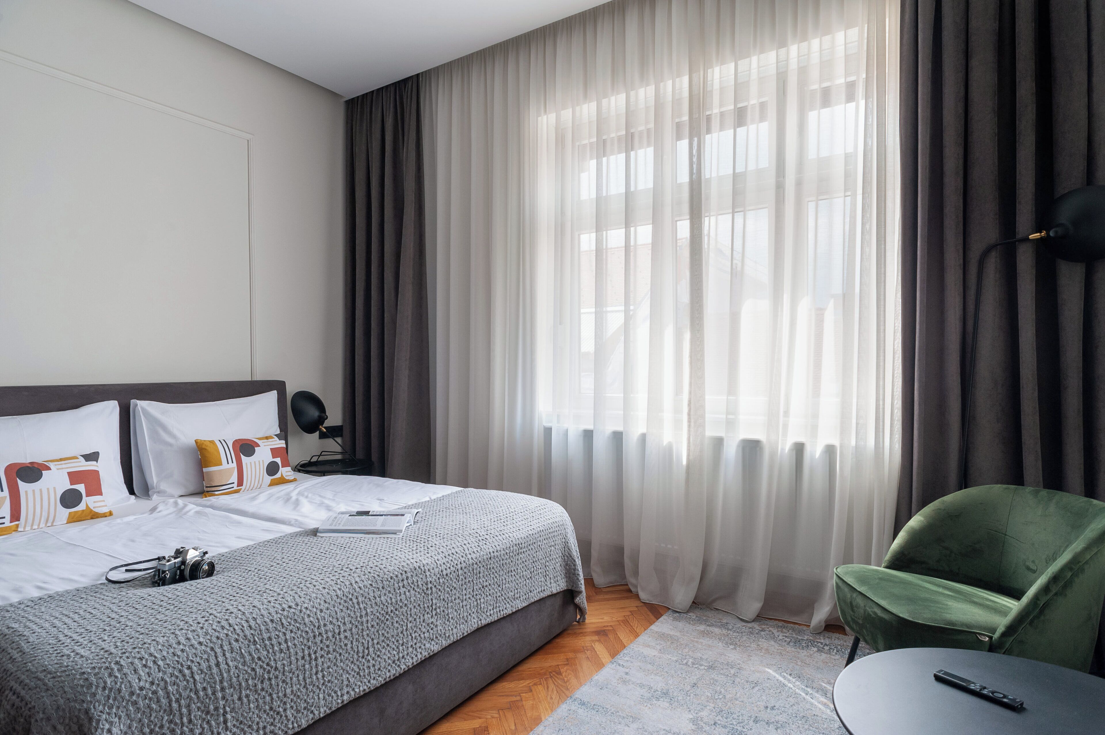 Een hotelkamer met een bed, een groene fauteuil, een tafeltje en een raam met dunne gordijnen.