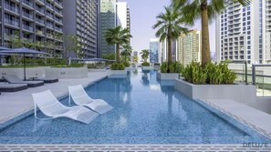 A modern hotel pool area with lounge chairs, palm trees, and high-rise buildings in the background.