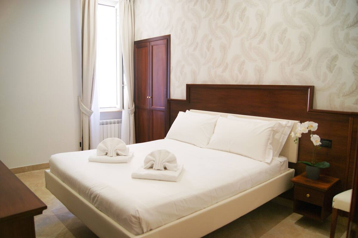 A neatly made bed with white linens and towels, a wooden headboard, a nightstand with a lamp, and a window with curtains.