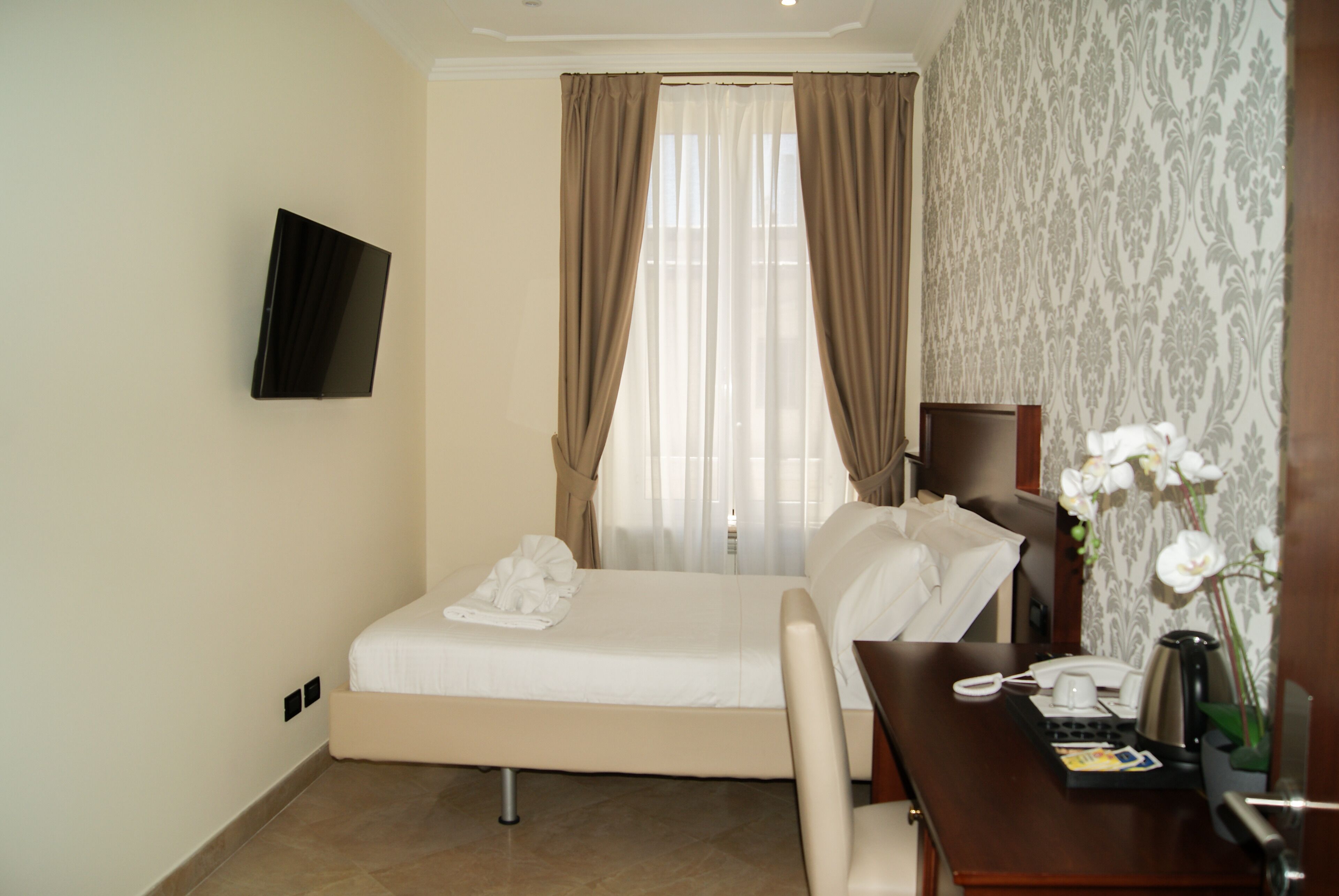 A hotel room with a bed, a desk with a telephone, a coffee maker, and a vase with flowers.