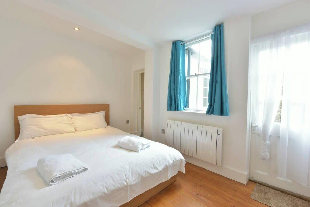 Una cama bien hecha con sábanas blancas y una toalla doblada, cabecera de madera, ventana con cortinas azules y un radiador blanco.