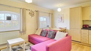 A cozy living room with a pink sofa, a white coffee table, and a kitchen area with wooden cabinets and a red appliance.