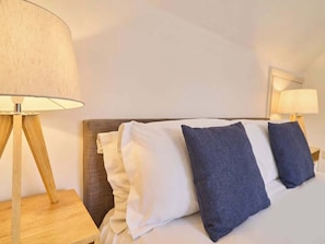 A neatly made bed with white and dark blue pillows, a wooden nightstand with a lamp, and a wall-mounted lamp. - Host & Stay | Queen's Corner (Canterbury)