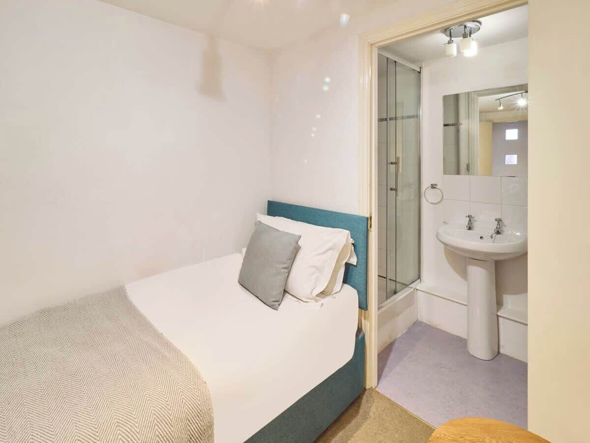 A bedroom with a bed, a bedside table, a mirror, and a bathroom with a sink and shower.