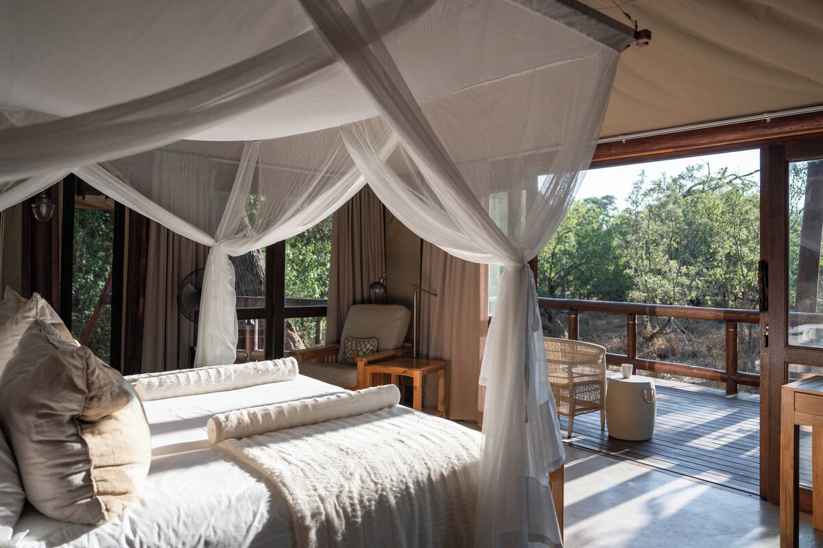 A bedroom with a canopy bed, a sitting area with a table and chairs, and a balcony with a view of trees.