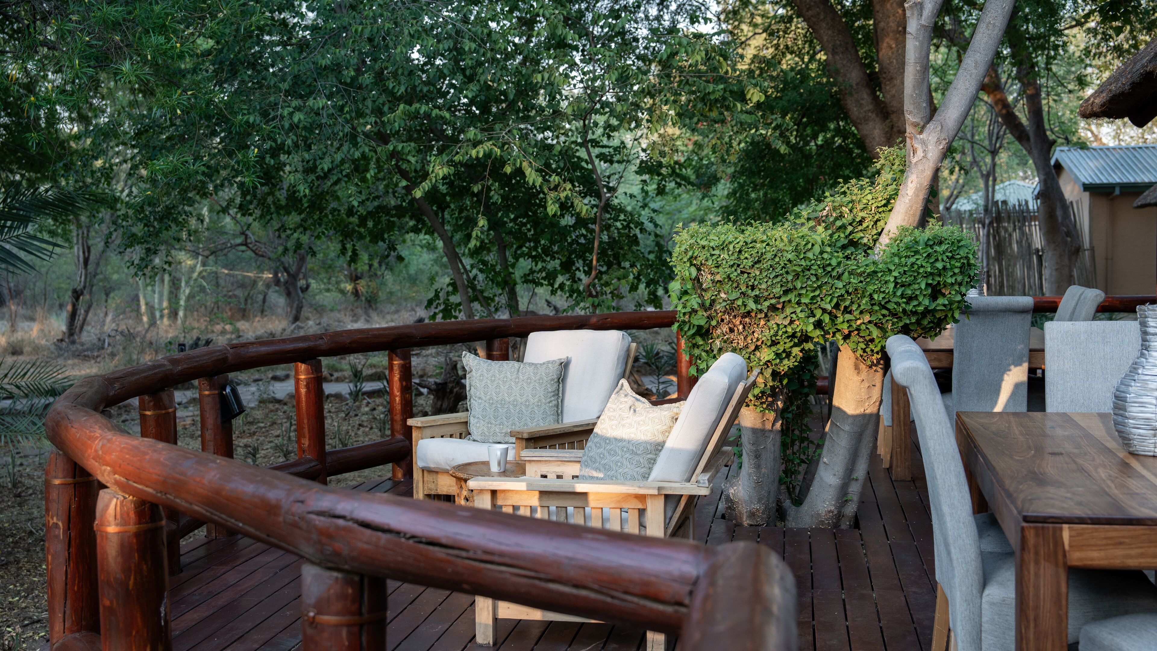 Una terraza de madera con mobiliario exterior, incluyendo sillas y una mesa, rodeada de exuberante follaje y árboles.