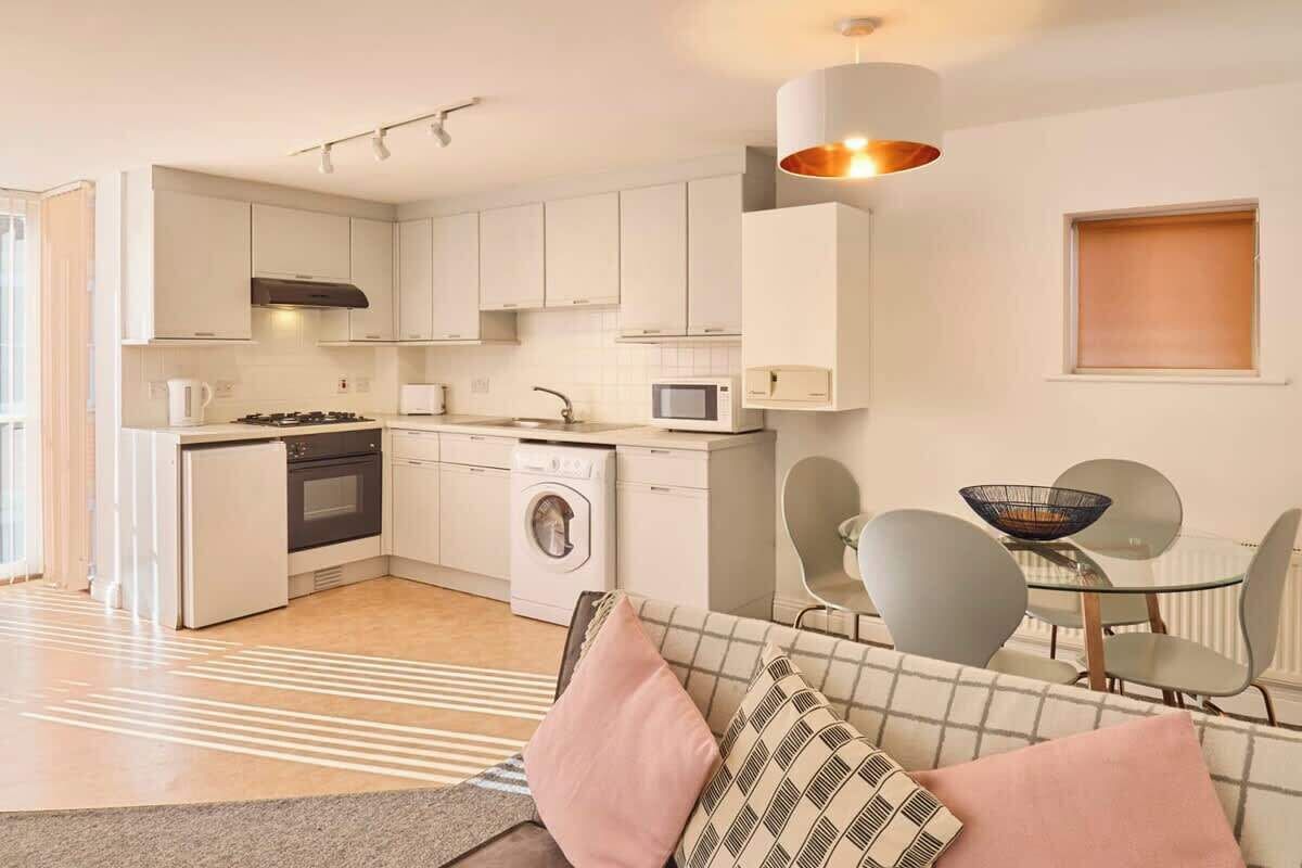 Ett modernt kök med vita skåp, en inbyggd ugn, mikrovågsugn och tvättmaskin. Det finns en matplats med ett glasbord och stolar, samt en soffa med mönstrade kuddar.