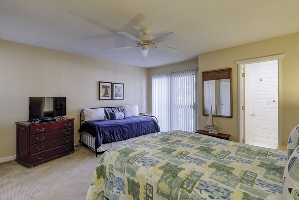 A bedroom with a bed, dresser, television, ceiling fan, window with blinds, and a door. - Sunset Rentals | Shipmaster 1703 | Dog Friendly (Hilton Head Island)