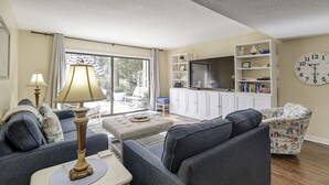 Un salon comprenant un canapé, un pouf, une lampe, un téléviseur, une bibliothèque et une horloge murale.