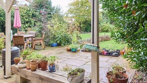 Ein überdachter Patio-Bereich mit Holzmöbeln, Topfpflanzen und Blick auf einen Garten.