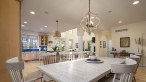 A spacious dining area with a large wooden table and white chairs, a hanging pendant light, and a kitchen in the background.