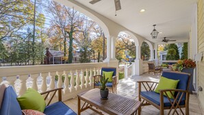 A covered outdoor patio with wooden furniture, a ceiling fan, and a view of trees and a distant building.