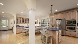 A spacious kitchen with wooden cabinets, a central island with stools, and a dining area with a large window.