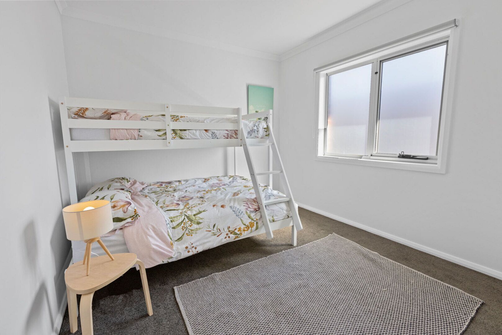 Un lit superposé avec une literie à motifs floraux, une petite chaise en bois, une lampe et une fenêtre avec des stores.