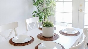 Ein Essbereich mit einem weißen Tisch, gedeckt für vier Personen, einer Hängelampe, einem Blumentopf und einem gerahmten Bild an der Wand.