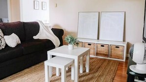 Ein Wohnzimmer mit einem dunkelgrauen Sofa, einem weißen Couchtisch und zwei weißen Beistelltischen. An der Wand hängen zwei gerahmte Kunstwerke, auf dem Couchtisch steht ein kleiner Blumentopf und auf dem Boden ein Korbwäschesack.