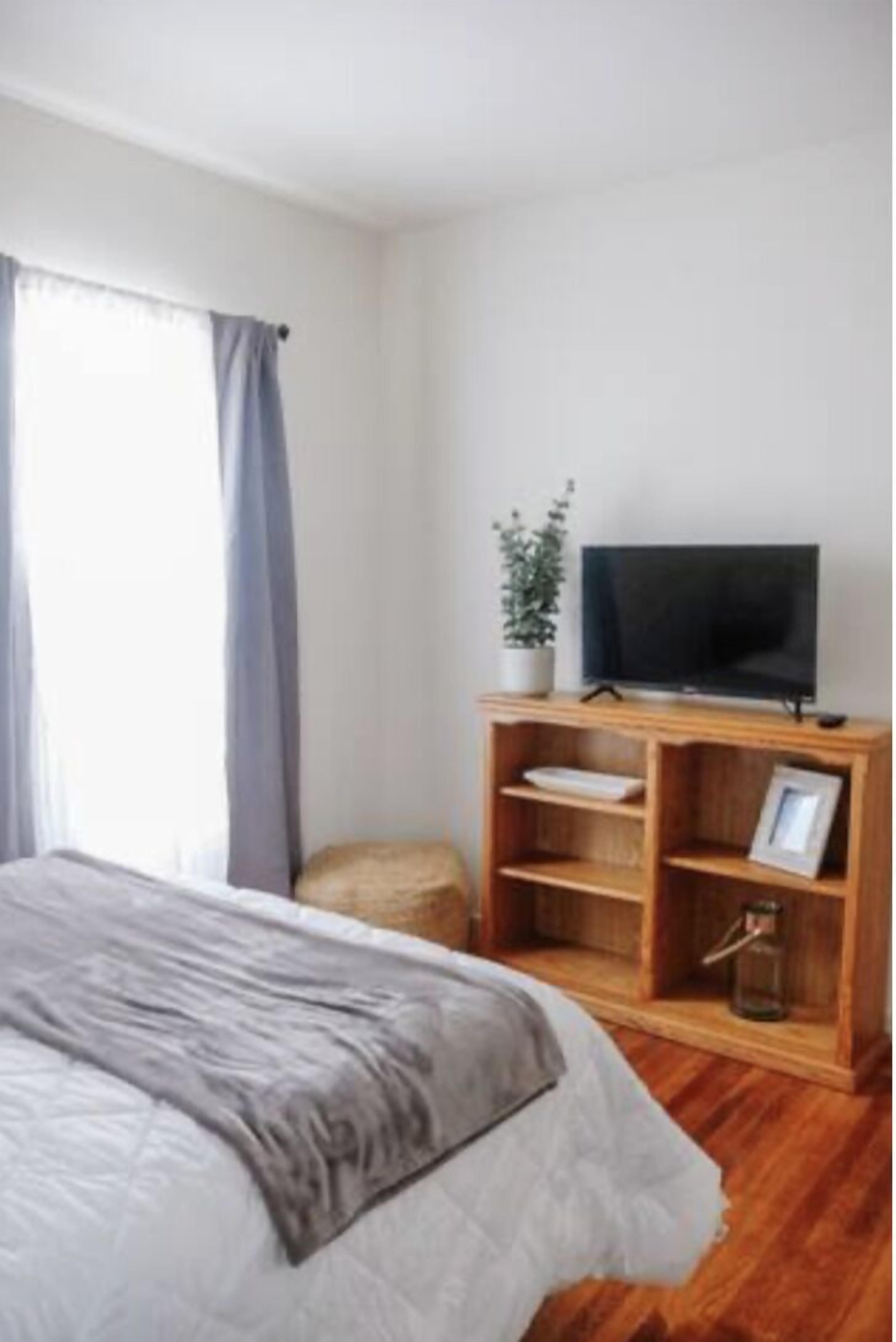 Ein Schlafzimmer mit einem Bett, einem Fernseher auf einem Holztisch, einer Topfpflanze und einem Fenster mit Vorhängen.