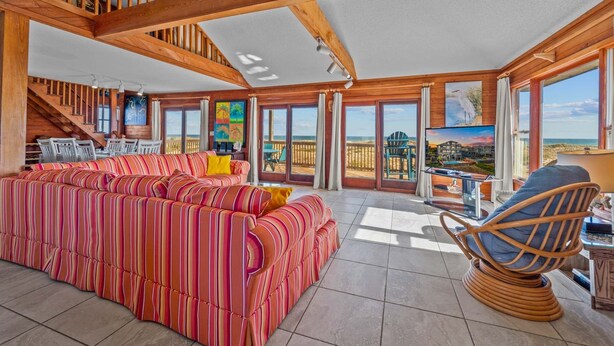 Un salon spacieux avec un grand canapé à rayures, un fauteuil en bois et un téléviseur. La pièce est dotée de grandes fenêtres qui offrent une vue sur la plage.