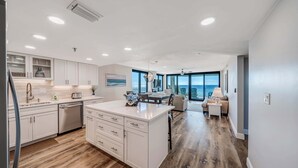 A modern kitchen with white cabinetry, a central island, and a dining area with a view. - Watercrest 302 Amazing VIEW, Pool, Hot Tub, Tennis, Gym, FREE Golf & Perks! (Panama City Beach)