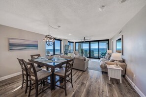 A spacious living area with a dining table, chairs, and a sofa. There are large windows offering a view of the ocean.