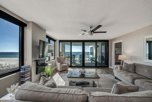 A spacious living room with a large window overlooking the beach, a ceiling fan, and a flat-screen TV mounted on the wall.