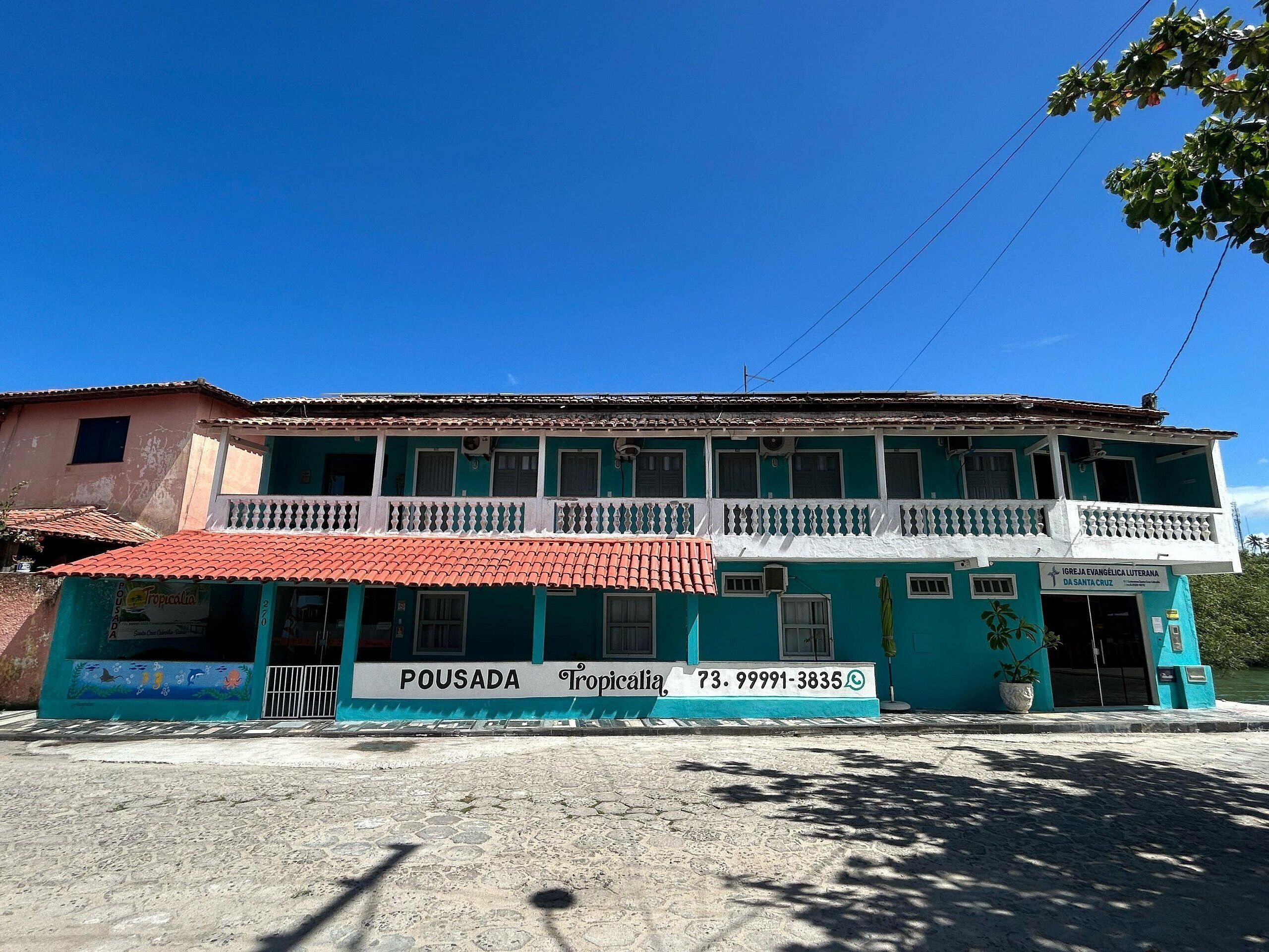 A two-story building with a balcony, a sign for 'POUSADA Tropicália,' and a contact number.