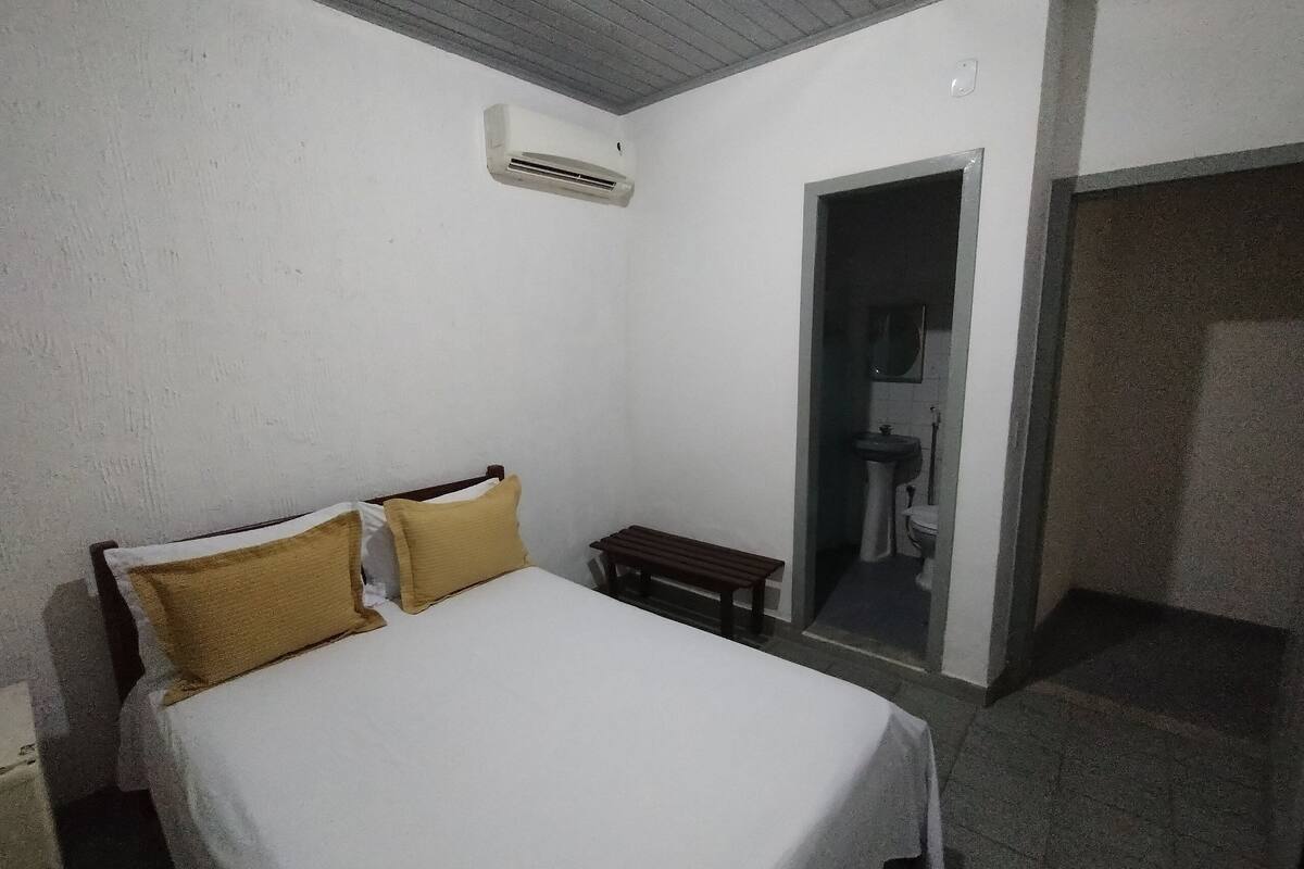 A bedroom with a bed, a bench, an air conditioning unit, and a bathroom door.