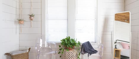 Una habitación con paredes de madera blanca, una silla de plástico transparente, un taburete tejido, una cama con cabecera de madera y dos ventanas con persianas.