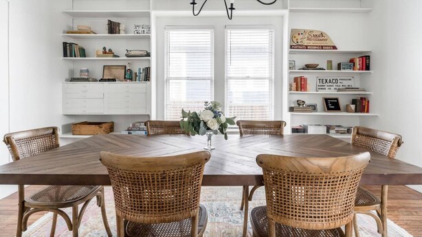 Ein Esszimmer mit einem großen Holztisch, sechs Korbstühlen, einer Chandelüster und weißen Regaleinbauten mit Büchern und Dekoration.