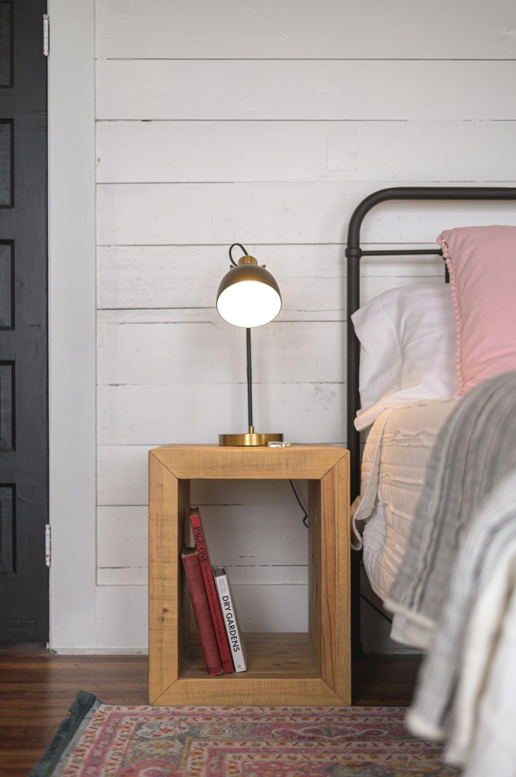 A wooden bedside table with a lamp, a bed with pink and white bedding, and a bookshelf with books.