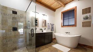 A bathroom with a glass shower enclosure, a freestanding bathtub, a vanity with dark cabinets, and a window with blinds. - Little Dutch Cabin 101-12 Min to Magnolia-baylor (Waco)