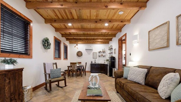 A cozy living room with a wooden ceiling, a brown sofa, a dining table with chairs, and a kitchen area in the background. - Little Dutch Cabin 101-12 Min to Magnolia-baylor (Waco)