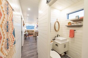 A narrow hallway leading to a bathroom with a toilet, sink, and shelves. There is a window with a view of the outside.