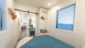 Une chambre à coucher avec un lit, une fenêtre avec des stores, une porte et un couloir menant à une autre pièce.